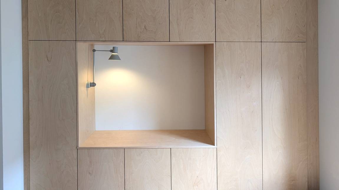 Plywood joinery wall with tight panel joints and a recessed seating nook lit by a small wall lamp