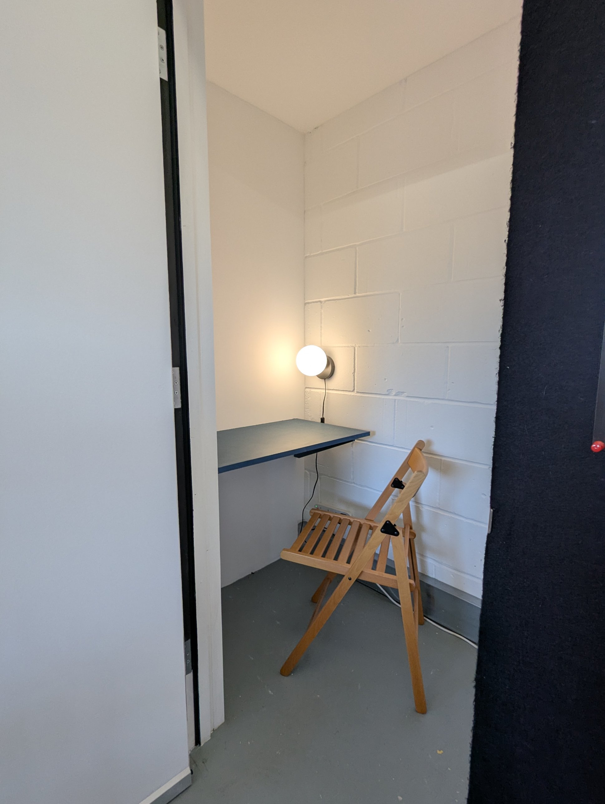 A small single-person booth with a narrow desk, wall light and folding chair, for taking calls away from the open floor